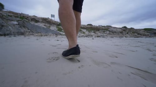Low-angled following shot of man's legs as he walks on beach towards exit stairs during overcast aft