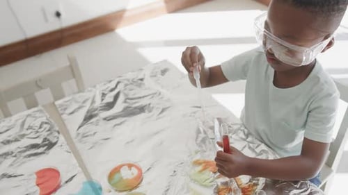 Niño afroamericano sentado en la mesa con tubos de ensayo con líquido, en cámara lenta