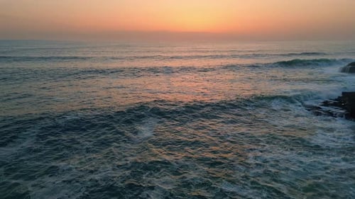 Pastel sunrise reflects on dramatic ocean waves splashing rocky cliffside aerial view