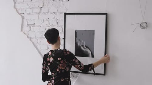 A Woman Gallerist Hangs New Paintings and Collages on the Wall of the Exhibition Hall