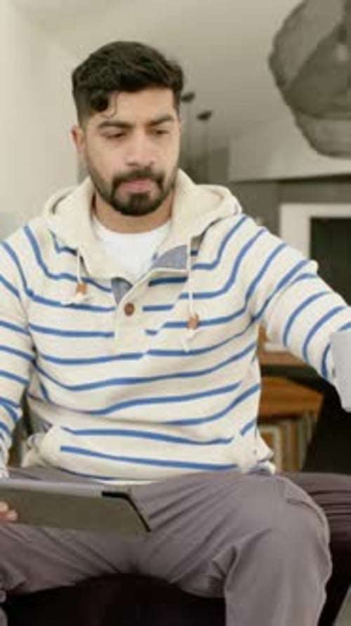 Vertical video: Bringing grey mug to lips, man in cream hoodie sipping while reading tablet at home