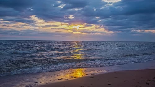Static shot of tropical waves crashing onto sandy beach during cloudy evening with sun setting in ti