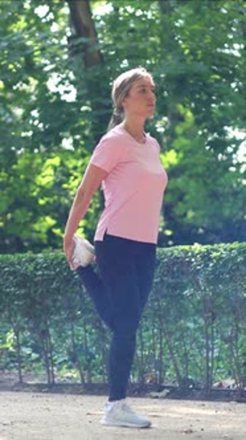 vertical Blonde female runner stretching leg muscles in a sunny park