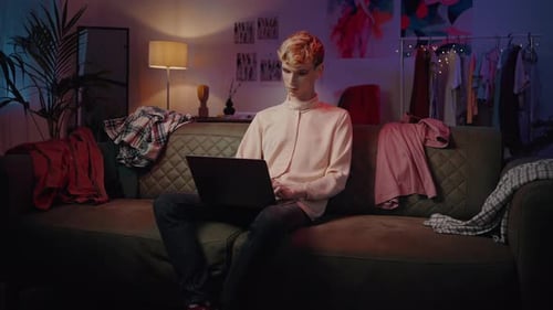 Young Nonbinary Guy Sitting on the Couch Working Remotely on Laptop at Home with Neon Lights