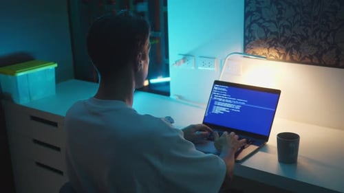 Programming at Night As Man Types Code on Laptop at Desk Under Blue Light Programming at Night