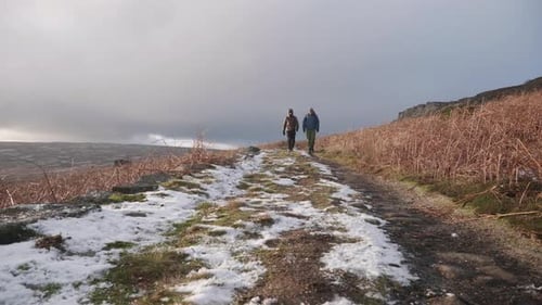 Two men hiking down a snowy, countryside trail towards the camera in beautiful winter sunshine and c
