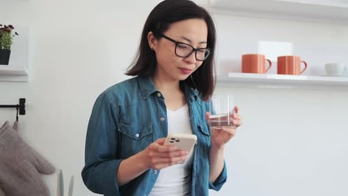 Woman Uses Phone, Drinks Water in Kitchen