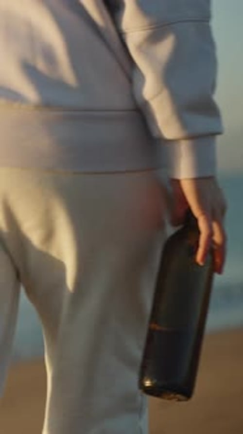 Vertical video. A lonely young woman warmly dressed with a bottle of wine Walks along the seashore