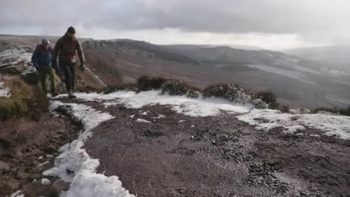 Two people hiking up a muddy, snowy winter path in the rural countryside with an epic view behind th