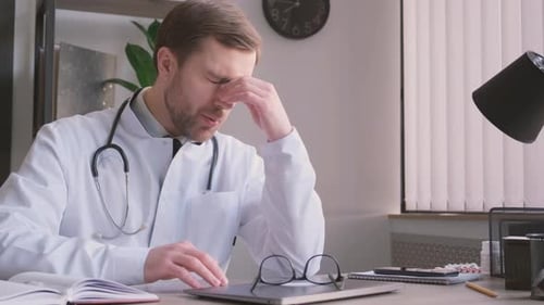 Handsome Young Doctor Sitting Alone in His Clinic and Feeling Stressed While Using His Laptop