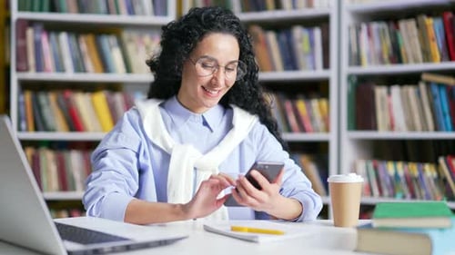 Smiling female student using smartphone sitting in campus library space. Happy girl looking
