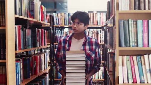 Young Male Student Walks Through Library Among Shelves with Books Holding Stack of Textbooks in
