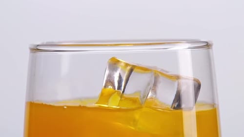 Close Up Of Ice Cube Falling In A Glass Of Orange Juice On The Light White Screen Background