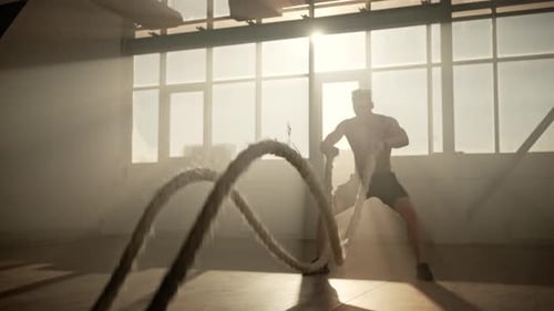 Muskulös aufgebauter Athlet, der in Innenräumen Herz-Kreislauf-Übungen mit Battle Ropes durchführt
