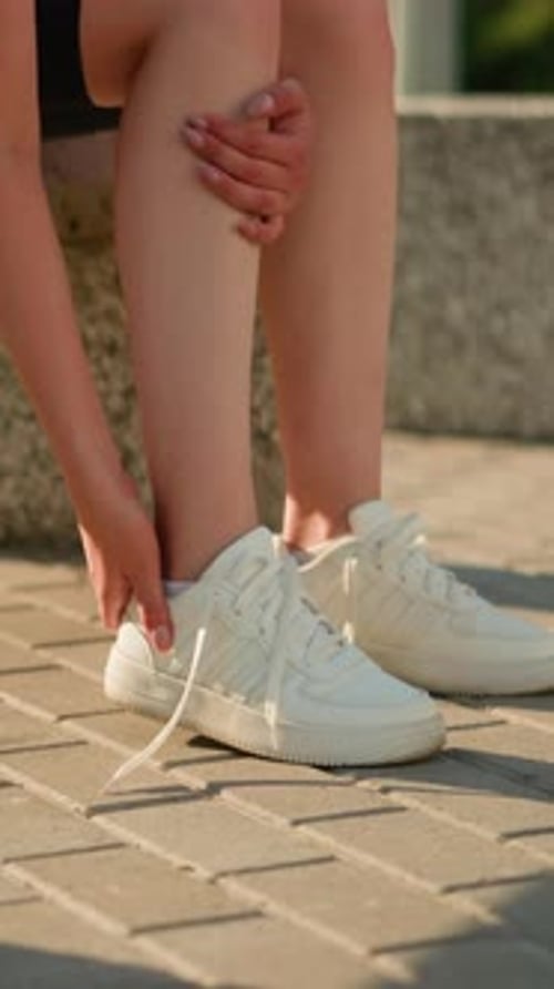 Lady Removing Sneaker Seated on Bench Outdoors with Greenery in Background