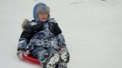 Slow motion of cheerful young boy sledging down a snowy hill on his plastic sleds