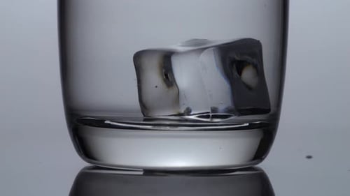 Close Up Of Ice Cube Falling In A Glass Of Drinking Water On The Light White Screen Background