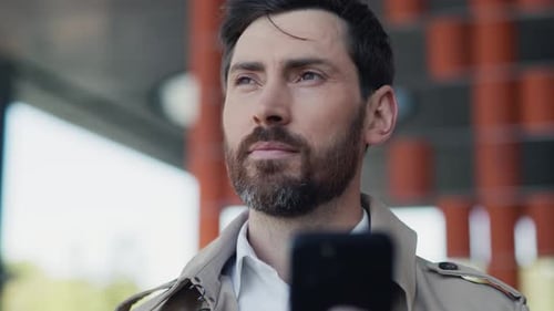 Attractive Young Stylish Handsome Man with Beard in Coat Uses Smartphone Stand in Part at Sunlight