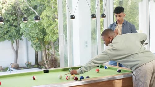 Diverse male friends striking cue ball at pool table scattering balls while preparing next shot