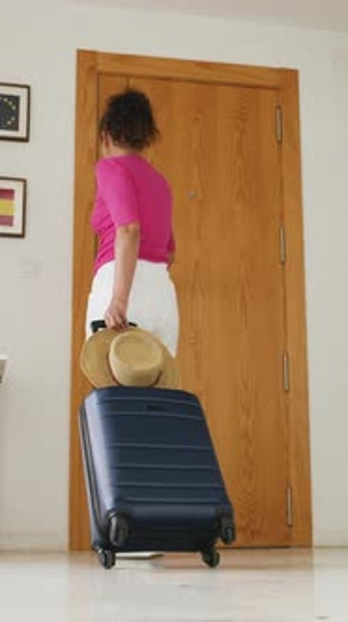 Mulher com mala azul escura e chapéu de sol está saindo do apartamento.