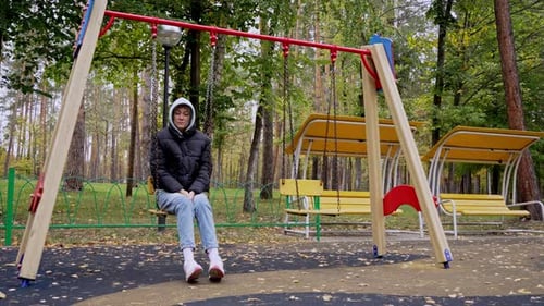 A pensive young woman in warm clothes sits on a swing in a city park