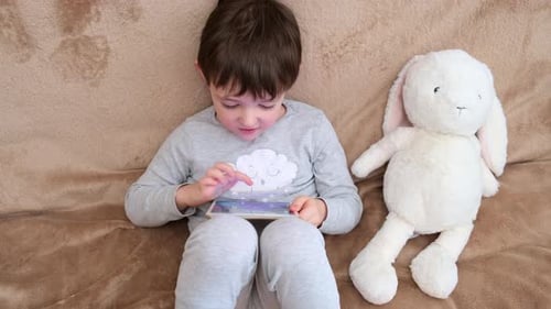 Child boy using tablet laying on sofa with stuffed toy. Kid enjoying digital content, screen time, g
