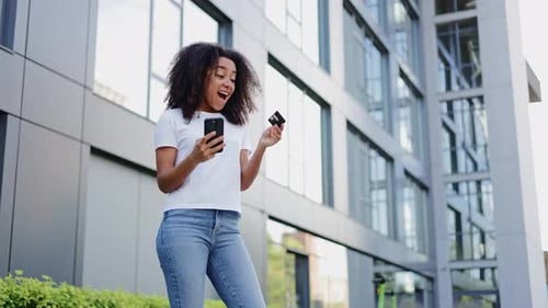 Cheerful Woman with Phone Pressing Debit Card to Chest While Smiling Outdoors