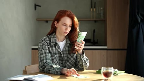 Portrait of Pensive Young Redhead Woman Holding Banknotes Feeling Stress About Unemployment at Home