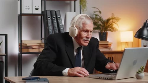 Overjoyed Senior Businessman Sitting at Home Office Desk Listening to Music Through Headphones
