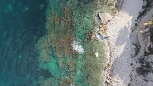 Rocky shoreline with crystal clear water meeting a small pebble beach, aerial view