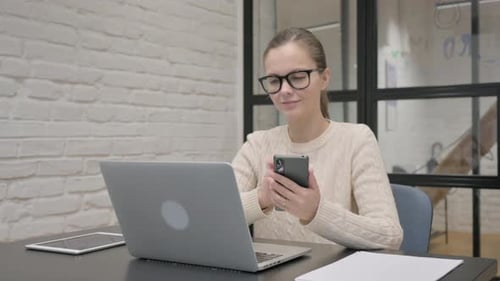 Woman Uses Phone at Desk in Front of Laptop