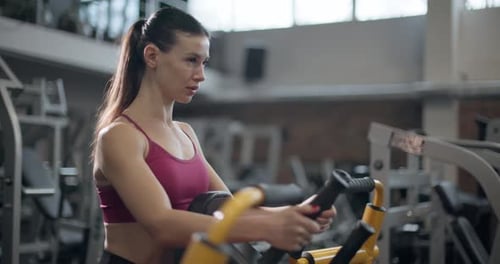 A Young Beautiful Brunette Girl in Sportswear is Exercising on a Machine in the Gym Doing Exercises