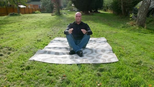 A man meditating in a yoga position in a garden. The mature male is sitting on a mat outside at home