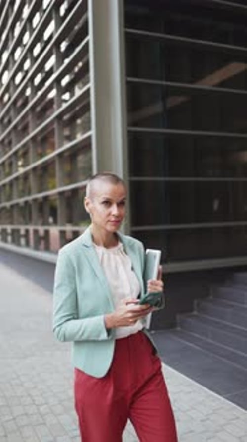 Vertical video of a modern business woman walking with shaved hair using mobile phone outside office