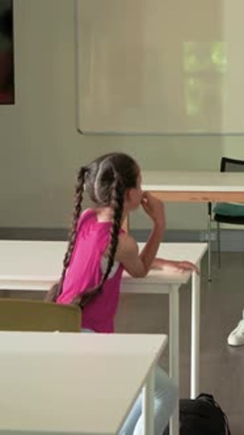 Vertical video: Turning to teacher entering, speaking, girl fiddling fingers at desk, backpack near