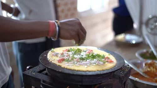Close-up shot of a man preparing Dosa, adding vegetables like onion on top with cooking oil in a sho