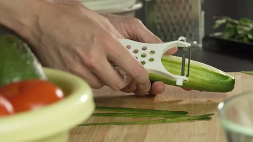 Hands of middle man european peels long cucumber with vegetable peeler to prepare salad, left side