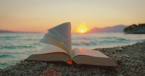Bibelseiten drehen sich am Kiesstrand auf See mit glühendem Sonnenuntergangslicht