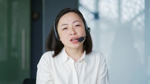 Webcam view. Asian woman in wireless headset customer service operator talking on video call while