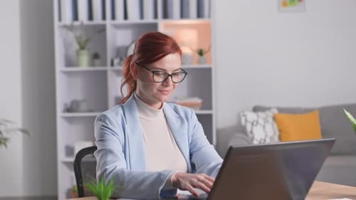 Porträt einer jungen Freiberuflerin mit Brille, die am Computer arbeitet, während sie lächelnd zu Hause sitzt