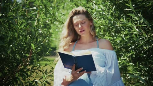 Woman Reading a Book in Lush Greenery on a Sunny Day By a Peaceful Garden