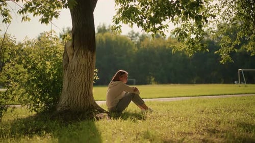 Woman Sitting By Tree in Sunlit Park Surrounded By Greenery