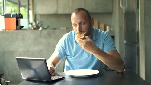 Man Eats Sandwich While Working on Laptop