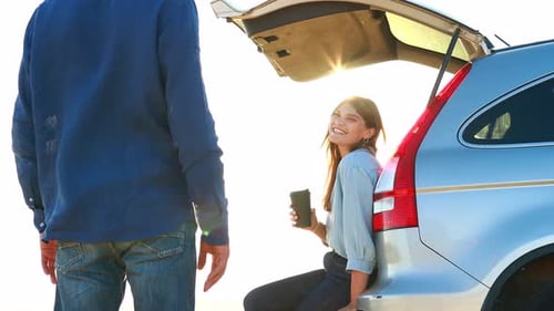 Happy Young Woman with Coffee Cup and Sitting on the Open Trunk of Her Car at Sunset and Laughing