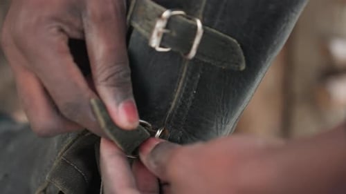 Petite boucle en cuir noire fermant les doigts sur une guêtre d'équitation, gros plan du fermoir en métal et des coutures