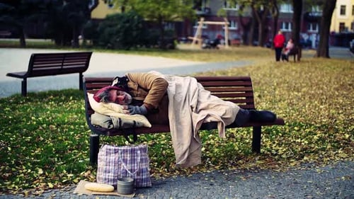 Homeless Beggar Man with a Pillow Lying on Bench Outdoors in City