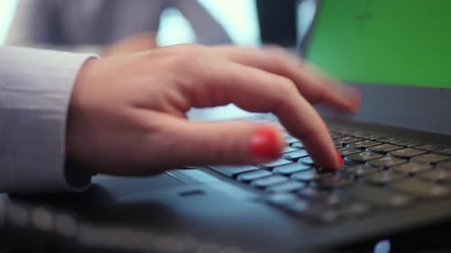 A female hand types a password on a laptop keyboard, dictated to her. The laptop screen has a green