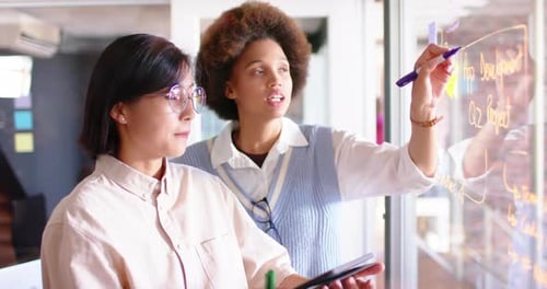Duas colegas criativas diversas discutindo ideias em uma parede de vidro, em câmera lenta
