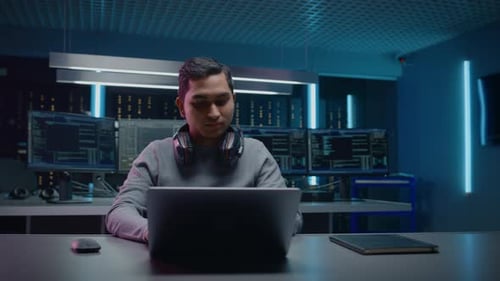 Portrait of Handsome Indian Software Developer / Hacker / Gamer Wearing Headset Sitting at His Desk