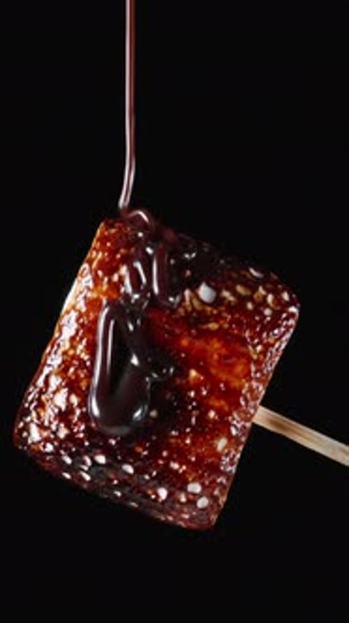 Toasted to a caramel crust, the marshmallow rotates and chocolate pours on it. Close-up on a black b